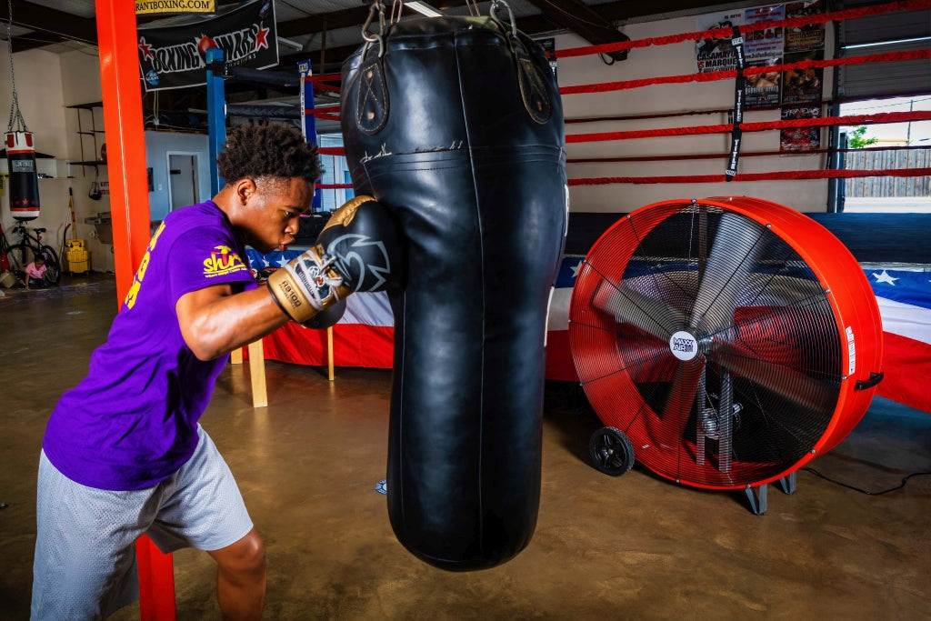 Boxer practices on the punching bag with a red Maxx Air drum fan cooling him near the ring. 