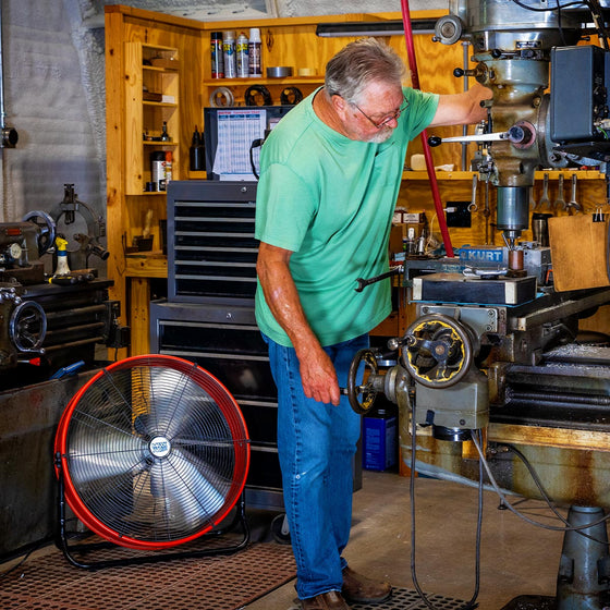 Employee in a factory works on machinery with the 24" tilt fan cooling him in the background. 